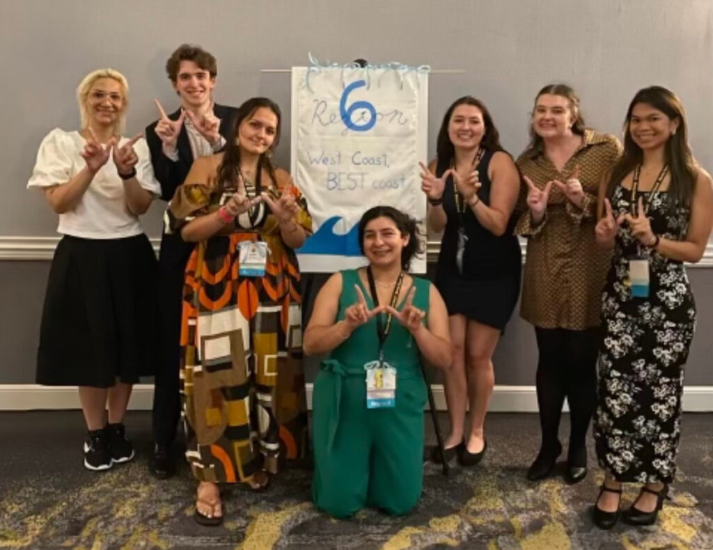 A group of eight Chapman Mortar Board members posing indoors in front of a handmade banner that reads ‘Region 6 – West Coast, BEST coast’ with a wave illustration. Everyone is smiling and holding up a ‘W’ hand sign. The group is dressed in a mix of formal and semi-formal attire.